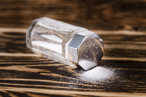 Some salt spilled out of the glass salt shaker with a silver metal lid lies on its side on a dark wooden table. Low key photography. Front view.
