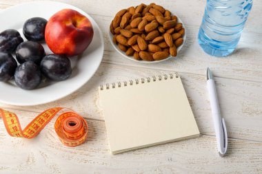 Kağıt not defteri, mezura, taze meyve, badem ve beyaz ahşap bir masada içme suyu. Defter sayfasına boşluk kopyala. Sağlıklı beslenme, diyet, vücut zayıflığı kavramı. Üst görünüm.