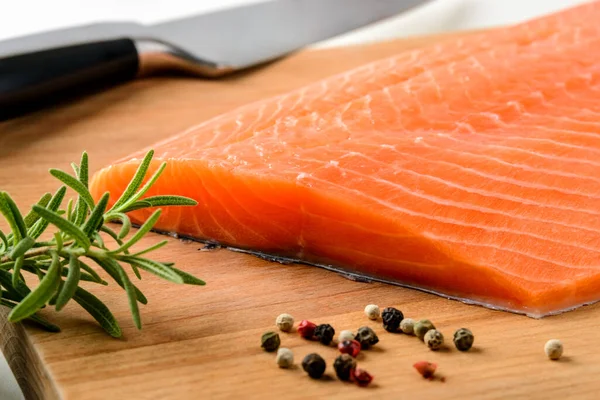 新鮮な生の鮭の切り身 鋭いナイフ 茶色の木のまな板にコショウの穀物とローズマリー 健康的な食事 魚介類のレシピ成分と有機オメガ3ソースの概念 正面図 — ストック写真