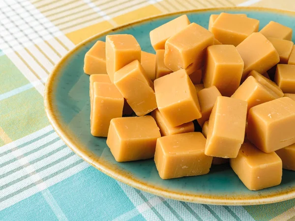 Caramel vanilla fudge candies on a turquoise ceramic plate over a checkered tablecloth. Fresh tasty chewy bonbons made of milk and sugar. Square pieces of delicious soft sweets. Top view.