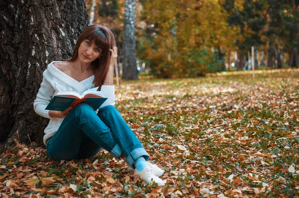 Piękna młoda brunetka siedzi na fallen jesieni liści w parku, czytanie książki — Zdjęcie stockowe