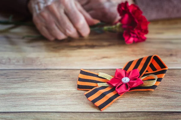 Concept background of May 9 Russian holiday Victory Day. St. Georges ribbon on a wooden table, old woman holding in hands a red carnation
