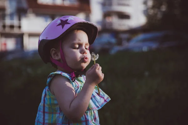 Fröhliches kleines Mädchen bläst Löwenzahnblüte in die Luft. Mädchen mit Helm — Stockfoto