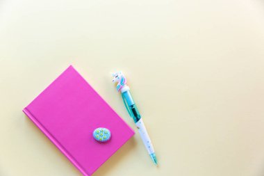 Notlar için pembe defter, komik tek boynuzlu at kalemi, pastel arka plan üzerine dekoratif yumurta. Düz yatıyordu. Üst te. Kopyalama alanı