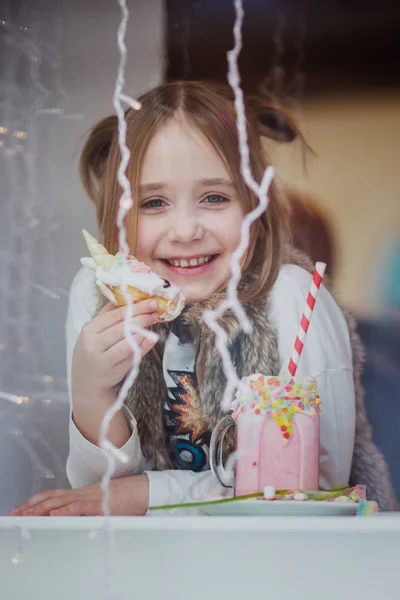 Симпатичная счастливая девушка в модной одежде пьет розовый молочный коктейль из трубки, украшенной пончиком в виде единорога, конфеты в кафе со светом на заднем плане — стоковое фото