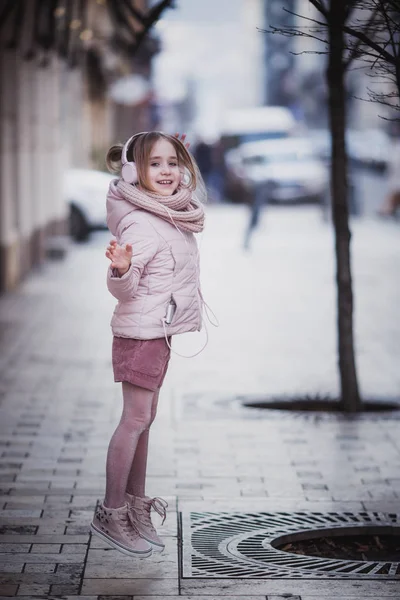 8-10 lat stary szczęśliwy dziewczyna w modne ciuchy słucha muzyki z jej smartphone w słuchawkach i skacze na zewnątrz. Słoneczny dzień w city — Zdjęcie stockowe