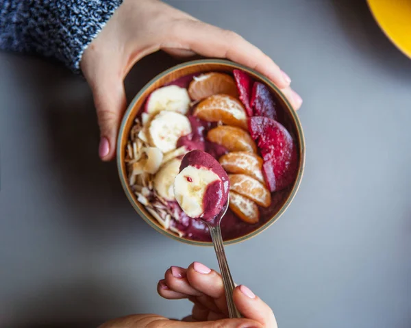 Acai smoothie, granola, seeds, fresh fruits in a wooden bowl in female hands on grey table. Eating healthy breakfast bowl. Top view — Stock Photo, Image