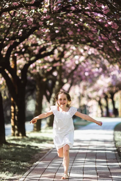 Entzückendes kleines Mädchen in weißem Kleid im blühenden rosa Garten an einem schönen Frühlingstag — Stockfoto