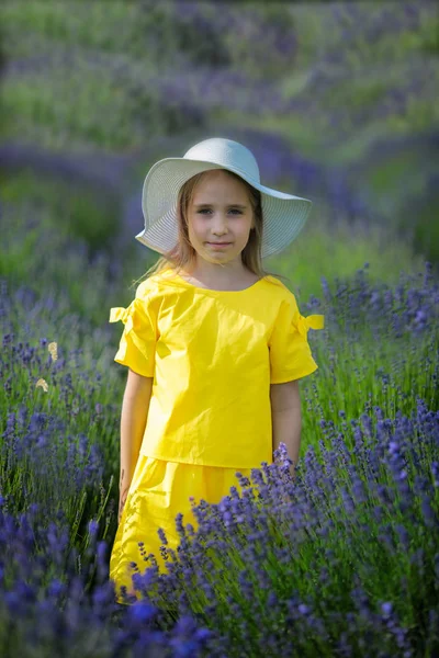 Schönes kleines Mädchen in einem gelben Kleid und weißem Hut, das in einem Lavendelfeld spaziert — Stockfoto