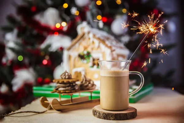 Nahaufnahme-Becher mit Kaffee und Milch auf einem Holztisch, Lebkuchenhaus und Weihnachtsbeleuchtung und Dekoration auf Bokeh. Wunderkerze brennt im Vordergrund — Stockfoto