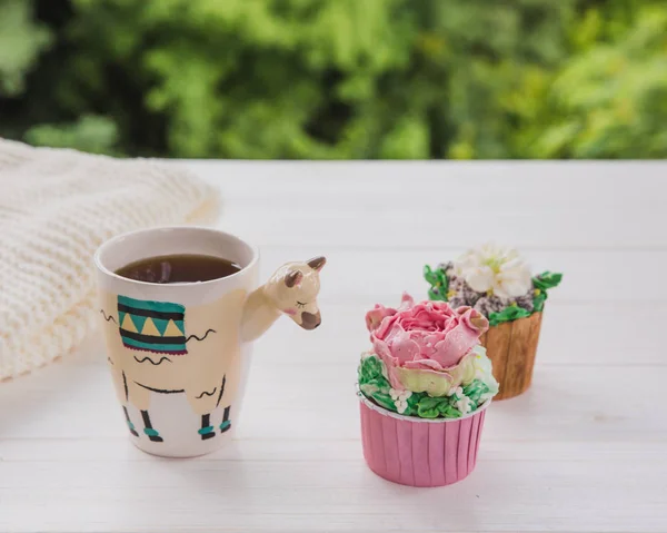 Hermosa taza de moda en forma de llama con bebida caliente y dos cupcakes en mesa de madera blanca con fondo verde brillante —  Fotos de Stock