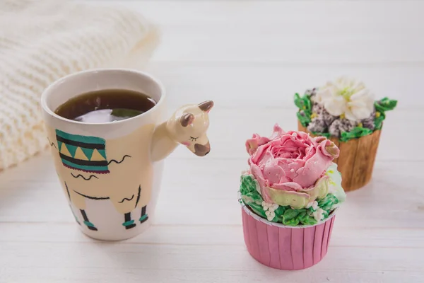 Hermosa taza de moda en forma de llama con bebida caliente y dos cupcakes en mesa de madera blanca con fondo verde brillante —  Fotos de Stock