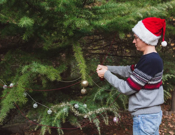 God jul och Happy Holidays. Far i Red Christmas hatt dekorera julgran utomhus på gården av huset före helgdagar — Stockfoto