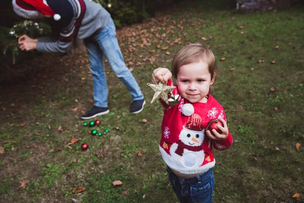 Merry Christmas and Happy Holidays. Little girl heps her father decorating the Christmas tree outdoor in the yard of the house before holidays — Stock Photo, Image
