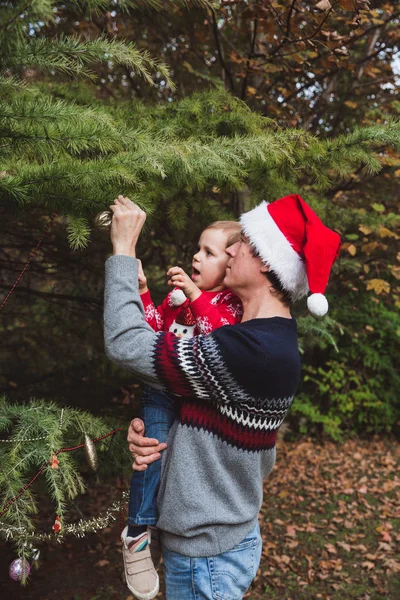 Счастливого Рождества и счастливых праздников. Отец в красной рождественской шляпе и дочь в красном свитере украшая елку на открытом воздухе во дворе дома перед праздниками — стоковое фото