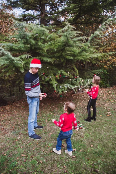 Wesołych Świąt i Wesołych Świąt. Ojciec w czerwonym kapeluszu Bożego Narodzenia i dwie córki w czerwone swetry dekorowanie choinki na zewnątrz w podwórku domu przed wakacjami — Zdjęcie stockowe