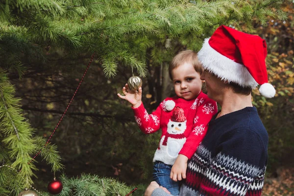 Merry Christmas and Happy Holidays. Father in red Christmas hat and daughter in red sweater decorating the Christmas tree outdoor in the yard of the house before holidays