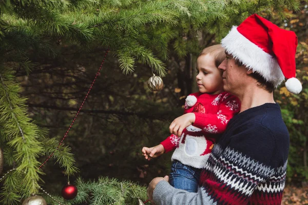 Счастливого Рождества и счастливых праздников. Отец в красной рождественской шляпе и дочь в красном свитере украшая елку на открытом воздухе во дворе дома перед праздниками — стоковое фото