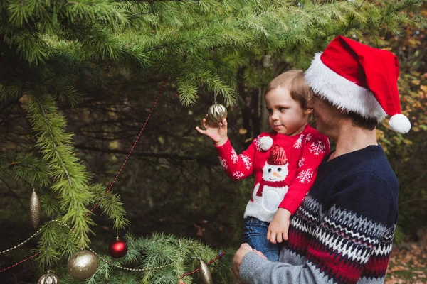 Merry Christmas and Happy Holidays. Father in red Christmas hat and daughter in red sweater decorating the Christmas tree outdoor in the yard of the house before holidays