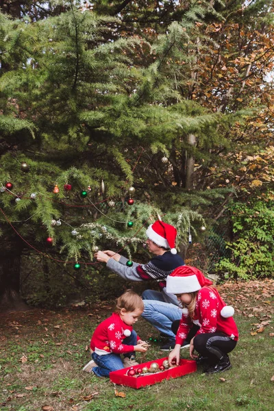 Merry Christmas and Happy Holidays. Father in red Christmas hat and two daughters in red sweaters decorating the Christmas tree outdoor in the yard of the house before holidays — Stock Photo, Image