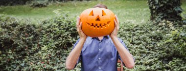 Mavi gömlekli adam yüzünün önünde büyük balkabağı tutuyor. Cadılar Bayramınız Kutlu Olsun