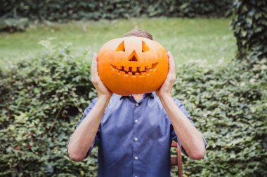 Mavi gömlekli adam yüzünün önünde büyük balkabağı tutuyor. Cadılar Bayramınız Kutlu Olsun