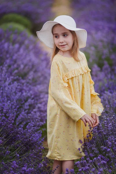 Uśmiechnięta dziewczynka w żółtej sukience i białym kapeluszu biegająca po polu lawendy — Zdjęcie stockowe