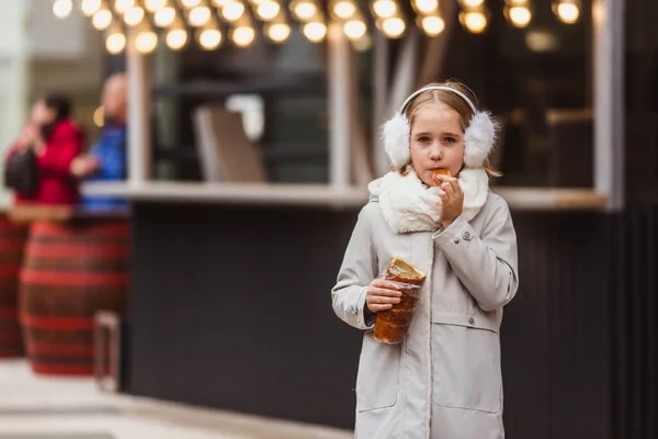 Маленькая милая девочка ест традиционное венгерское сладкое печенье под названием Kurtoskalacs в Будапеште на уличном рынке зимой — стоковое фото