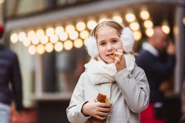 Маленькая милая девочка ест традиционное венгерское сладкое печенье под названием Kurtoskalacs в Будапеште на уличном рынке зимой — стоковое фото