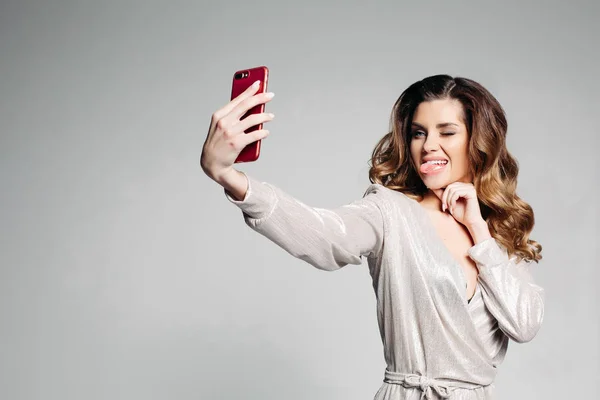 Portrait of a brunette woman dressed in a stylish gray dress, with make-up and hairstyle, makes selfie showing her tongue and fisting her eyes, for social networks. — Stock Photo, Image