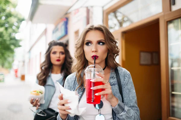 Belle ragazze con trucco e acconciatura bere bevanda frizzante e mangiare fast food . — Foto Stock