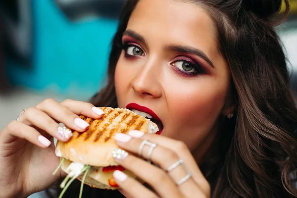 Dziewczyna z jasnym makijażu biorąc gryza hamburgera. — Zdjęcie stockowe