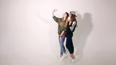 Güzel ve şık sarışın ve esmer kızlar, eğleniyor, birlikte fotoğraf yapma ve stüdyoda beyaz arka plan üzerinde poz. Kendi portresini çekmek konusunda yağma kız şık giyim, telefon.