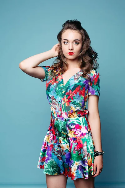 Ganska härlig tjej med vågigt hår i vacker blommig klänning poserar över Blå bakgrund. — Stockfoto