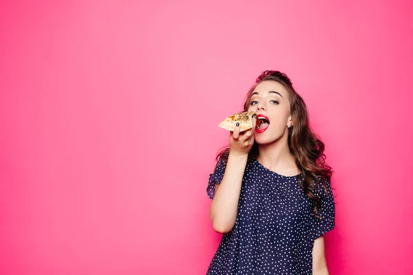Happy girl eating a pizza. She opened her mouth, holding a pizza in her right hand and staring at you with satisfaction. Isolated on pink background — Stock Photo, Image