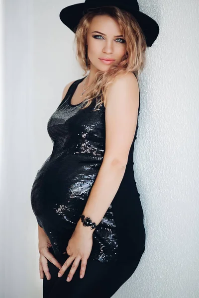 Hermosa mujer rubia rizada embarazada en vestido negro brillante y sombrero . —  Fotos de Stock