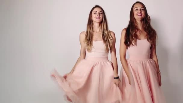 Vista frontal de dos chicas atractivas y elegantes agitando sus faldas acampanadas, bailando y saltando. Adorables chicas jóvenes con vestidos de color rosa pastel de moda, sonriendo, lanzando besos a la cámara . — Vídeos de Stock