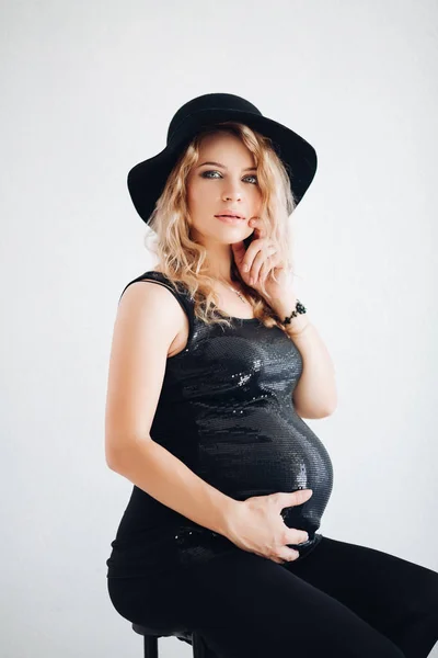 Mujer embarazada con estilo en sombrero negro y la parte superior brillante abrazando su vientre . —  Fotos de Stock