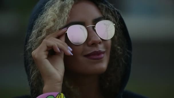 Hipster-Mädchen mit lockigem blondem Haar und verspiegelter Sonnenbrille, die wegschaut und lächelt. — Stockvideo