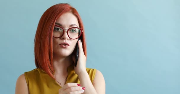 Röd haired kvinna i glasögon pratar på Ring samtal på blå bakgrund. — Stockvideo