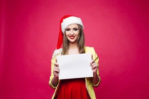 在圣诞节帽子和黄色外套的幸福女孩的肖像在红色的礼服持有白色空白的一张纸在明亮的粉红色背景 — 图库照片