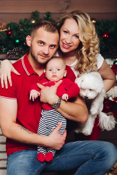 Family posing together near fireplace holding on hands little son and white puppy.