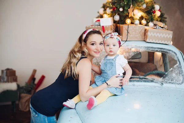 Fashionable blonde mom with little cute daughter posing together, sitting on blue retro car.