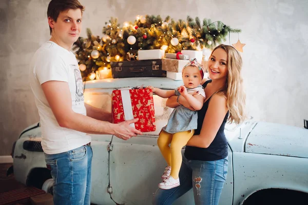 Precioso papá dando regalo de Navidad, caja roja grande, para la mamá rubia y la pequeña hija linda . —  Fotos de Stock