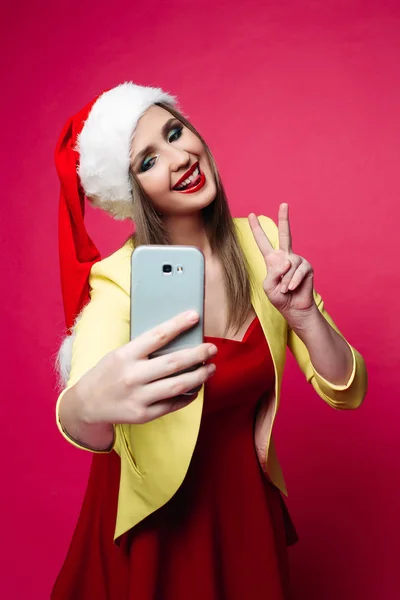 漂亮的白种人妇女穿着黄色夹克 红色的礼服和圣诞老人的帽子拍摄自肖像通过手机在生动的粉红色背景的工作室肖像 — 图库照片