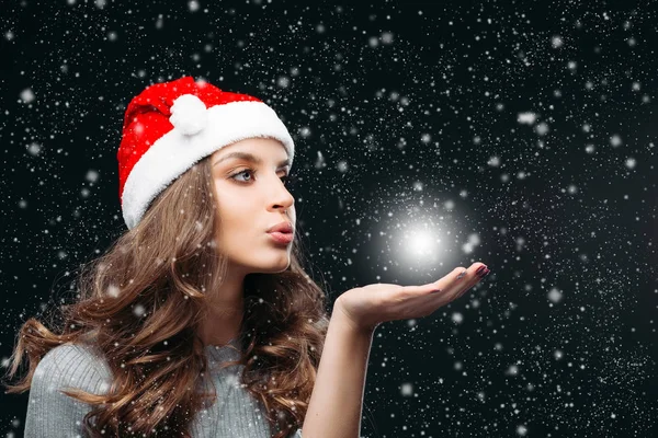 산타 모자를 쓴 아름다운 소녀가 눈이 내리는 동안 손으로 불고 있다. — 스톡 사진