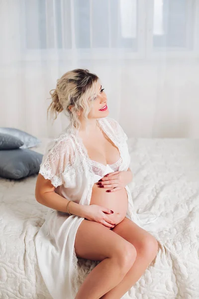 Sorrindo mulher muito grávida em lingerie branca na cama . — Fotografia de Stock