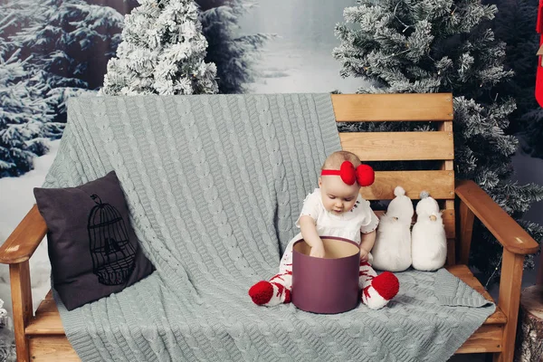 Bebek güzel kız şirin elbise ile lots-in Noel bankta oturmuş kafa bandı ile gülümseyen sunar. — Stok fotoğraf