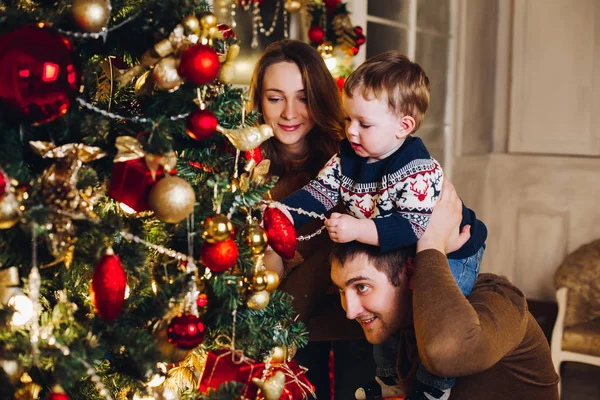 어린 아들을 위해 크리스마스 장식을 나무에 걸어 놓은 행복 한 부모들. — 스톡 사진