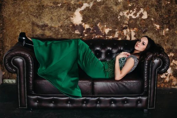 黒革のソファに横になっている緑のドレスでゴージャスで美しい女性. — ストック写真